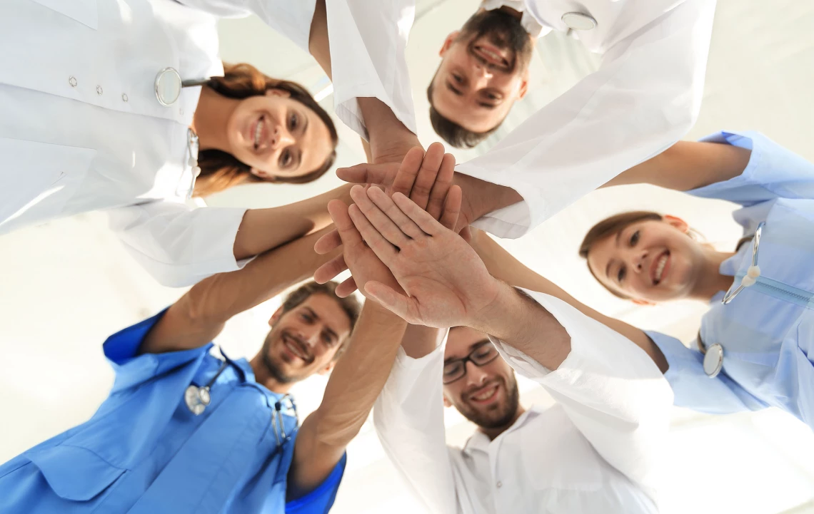 Fünf junge Ärzte stehen im Kreis über die Kamera gebeugt und halten die Hände zusammen.