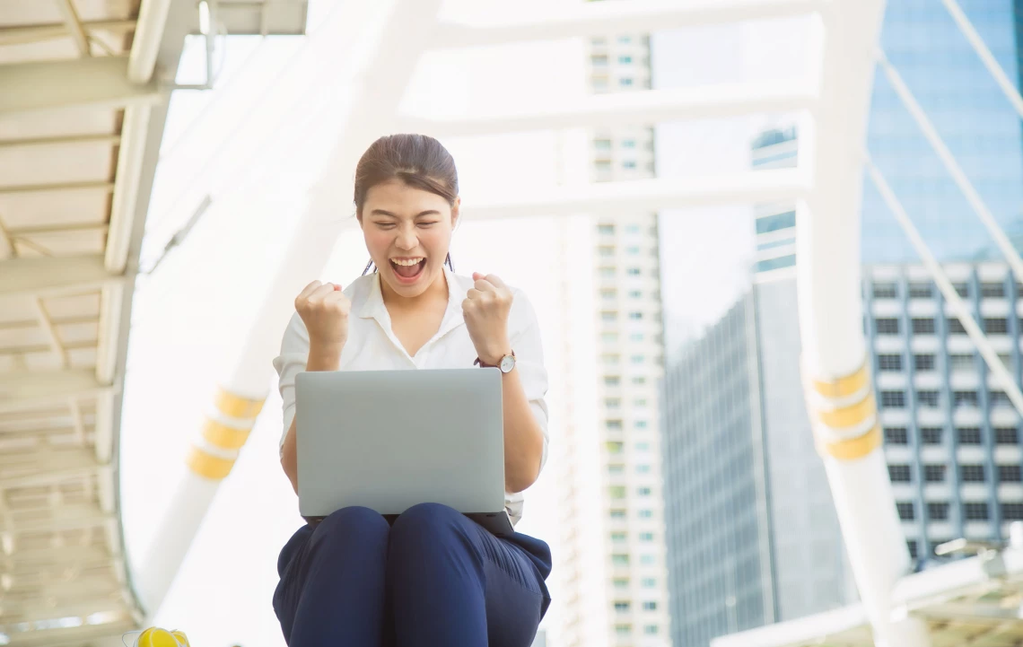 Eine Frau freut sich das sie angenommen wurde. Sie sitzt auf einer Treppe und schaut auf einen Laptop.