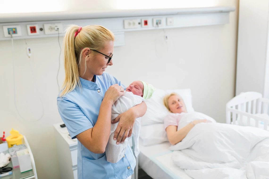 Eine Krankenschwester hält ein Neugeborenes. Daneben im Bett dessen Mutter.