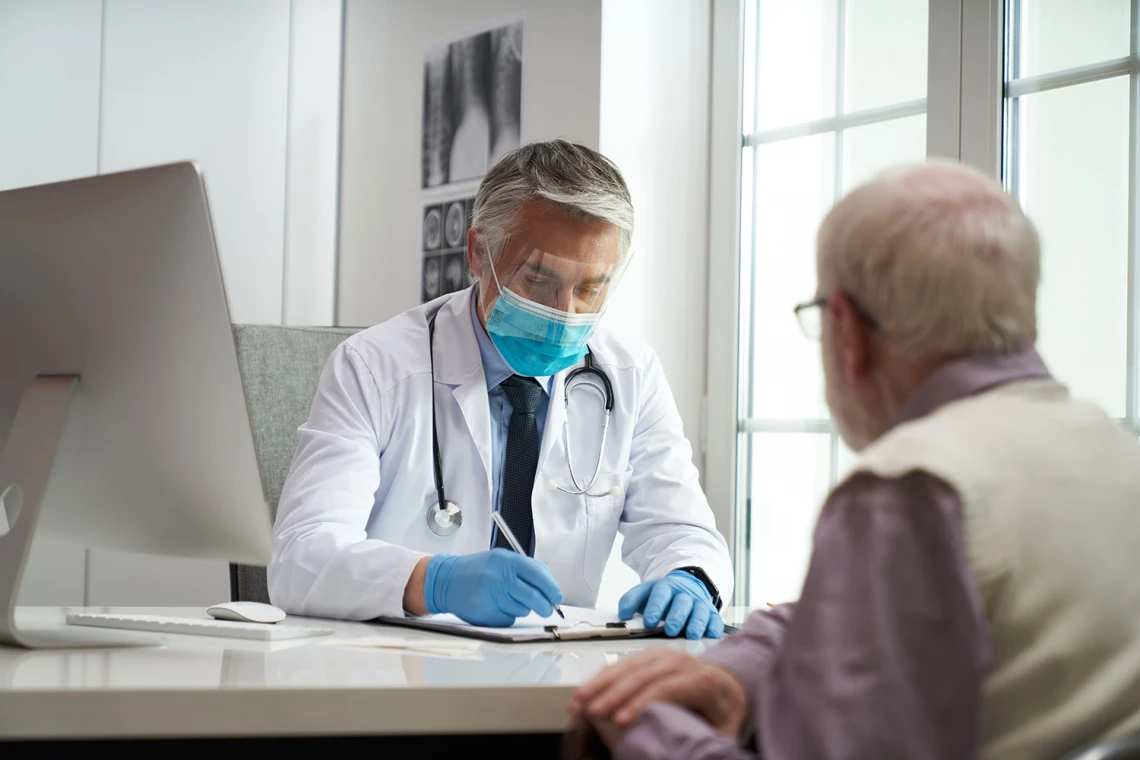 Ein Arzt redet mit einem Patienten während er auf eiem Klemmbrett schreibt.
