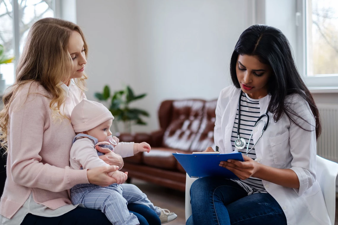 EIne Ärztin wertet zusammen mit einer Mutter die Untersuchung ihres Babys aus. Dieses sitzt auf dem Schoß der Mutter.