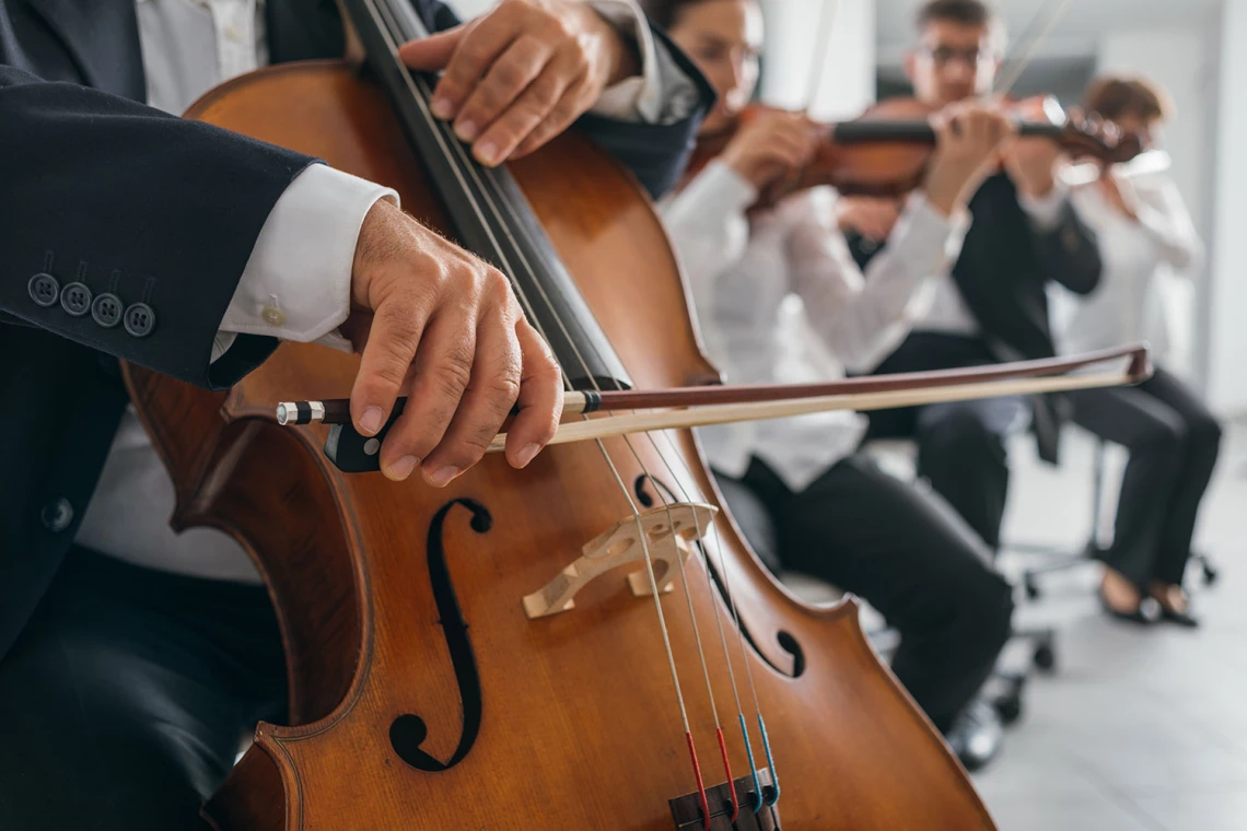 Ein kleines Orchester spielt in einem Saal. Im Vordergrund sieht man eine Nahaufnahme eines Cellos.