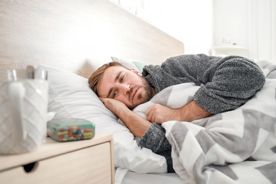 Ein Mann liegt krank im Bett. Auf seinem Nachttischschrank liegen Tabletten.