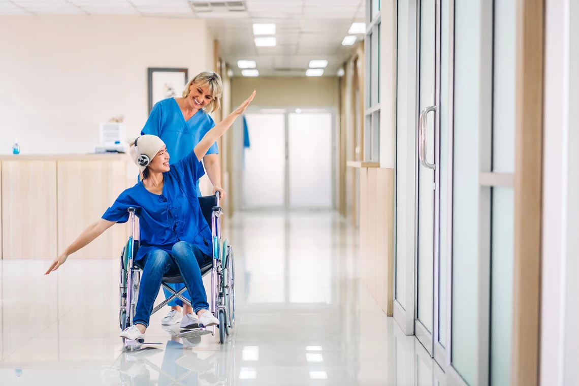 Eine Patientin im Rollstuhl imitiert ein Flugzeug und wird dabei von einer Krankenschwester geschoben.