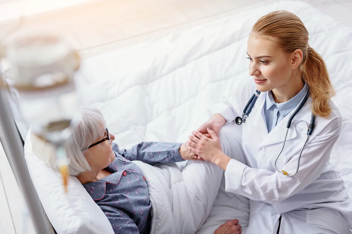 Eine rztin hält die Hand einer Patientin die im Bett liegt und hört sich ihr Anliegen an.